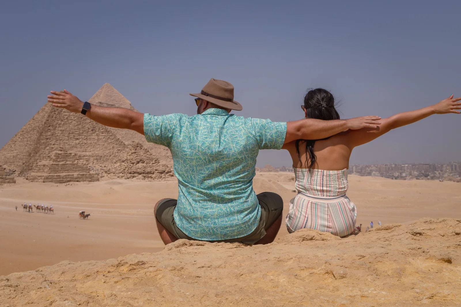 A couple sitting on a sand ridge with arms outstretched, facing the Pyramids of Giza