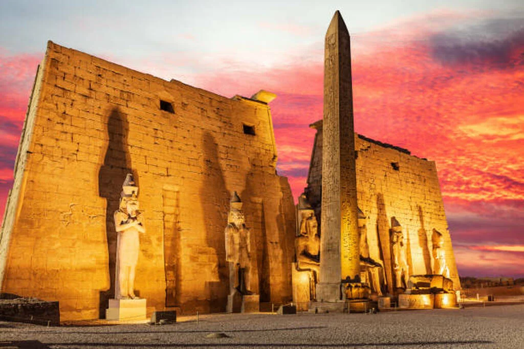The floodlit first pylon and obelisk of Luxor Temple under a red evening sky