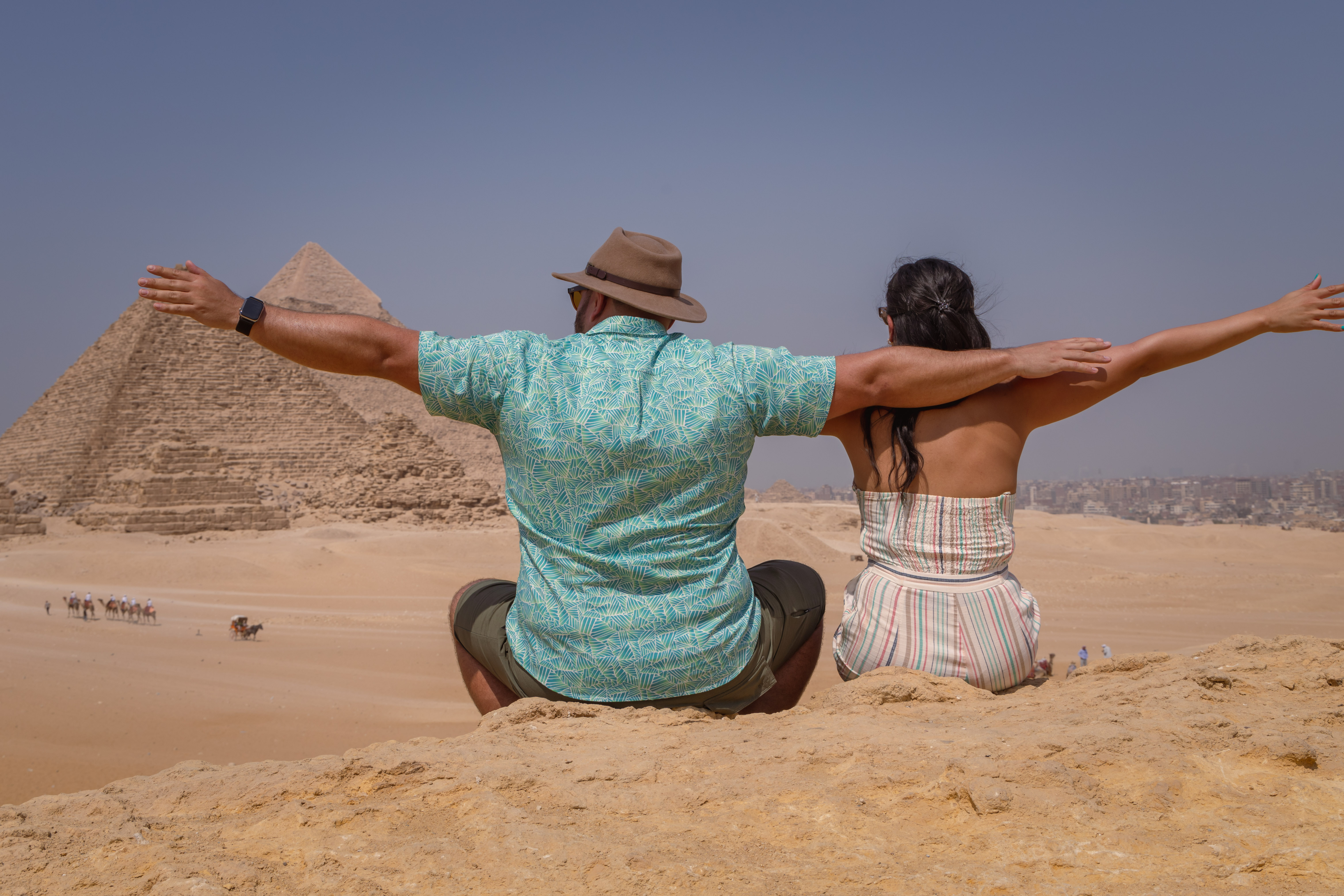 Two travellers sitting on the sand with arms outstretched in front of the Pyramids of Giza near Cairo